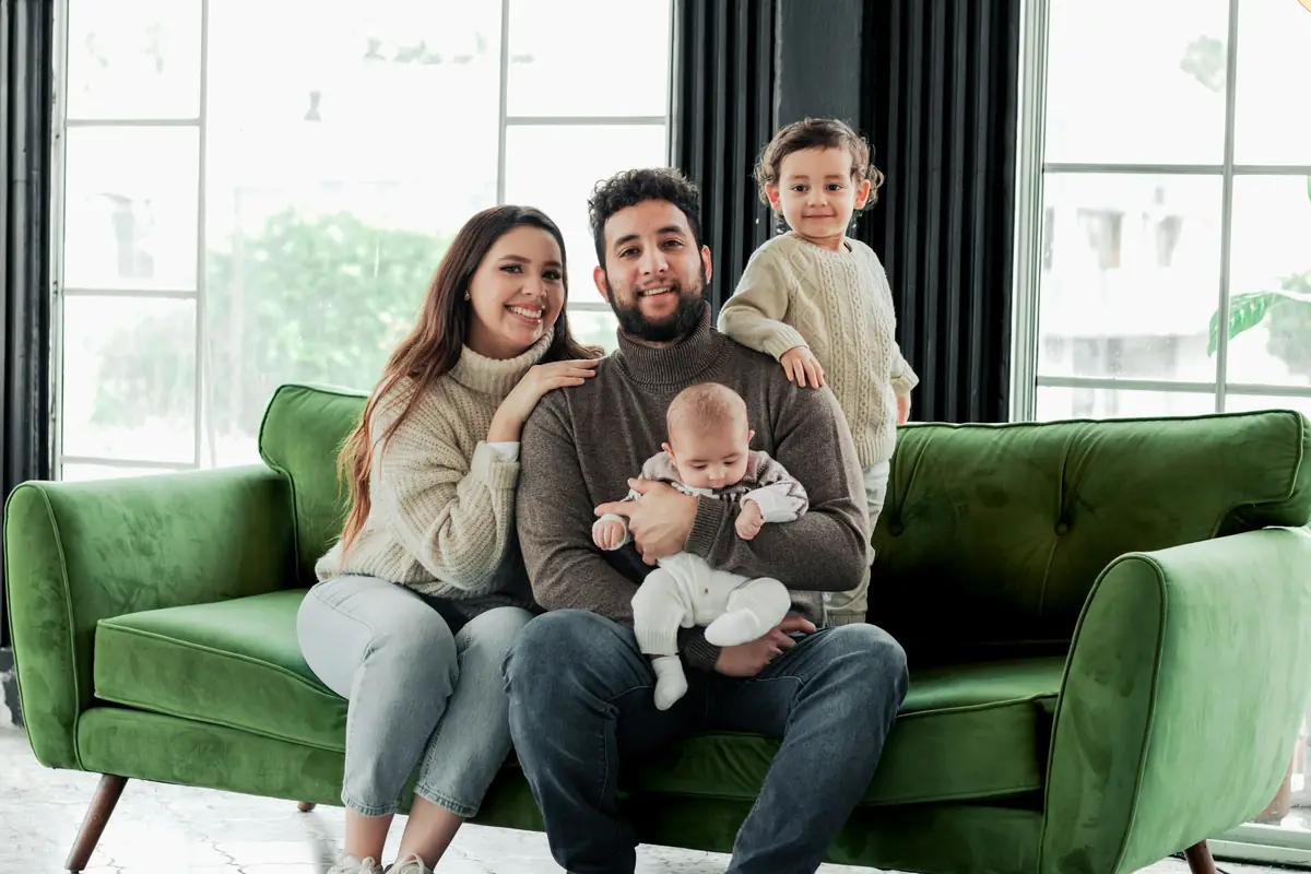 A family with a baby and a young child on a green sofa in a bright living room.
